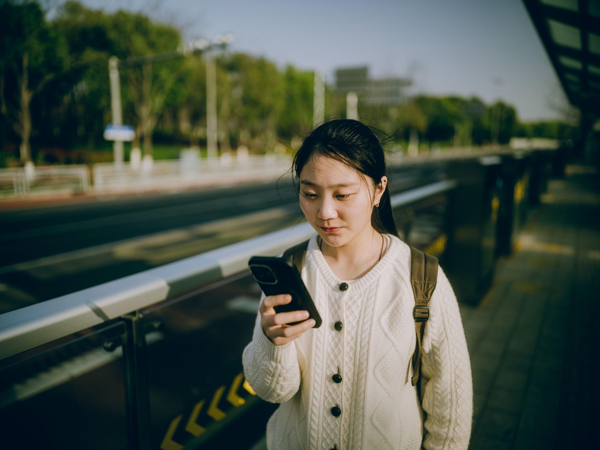 A young adult checks a smartphone while waiting at a train platform.